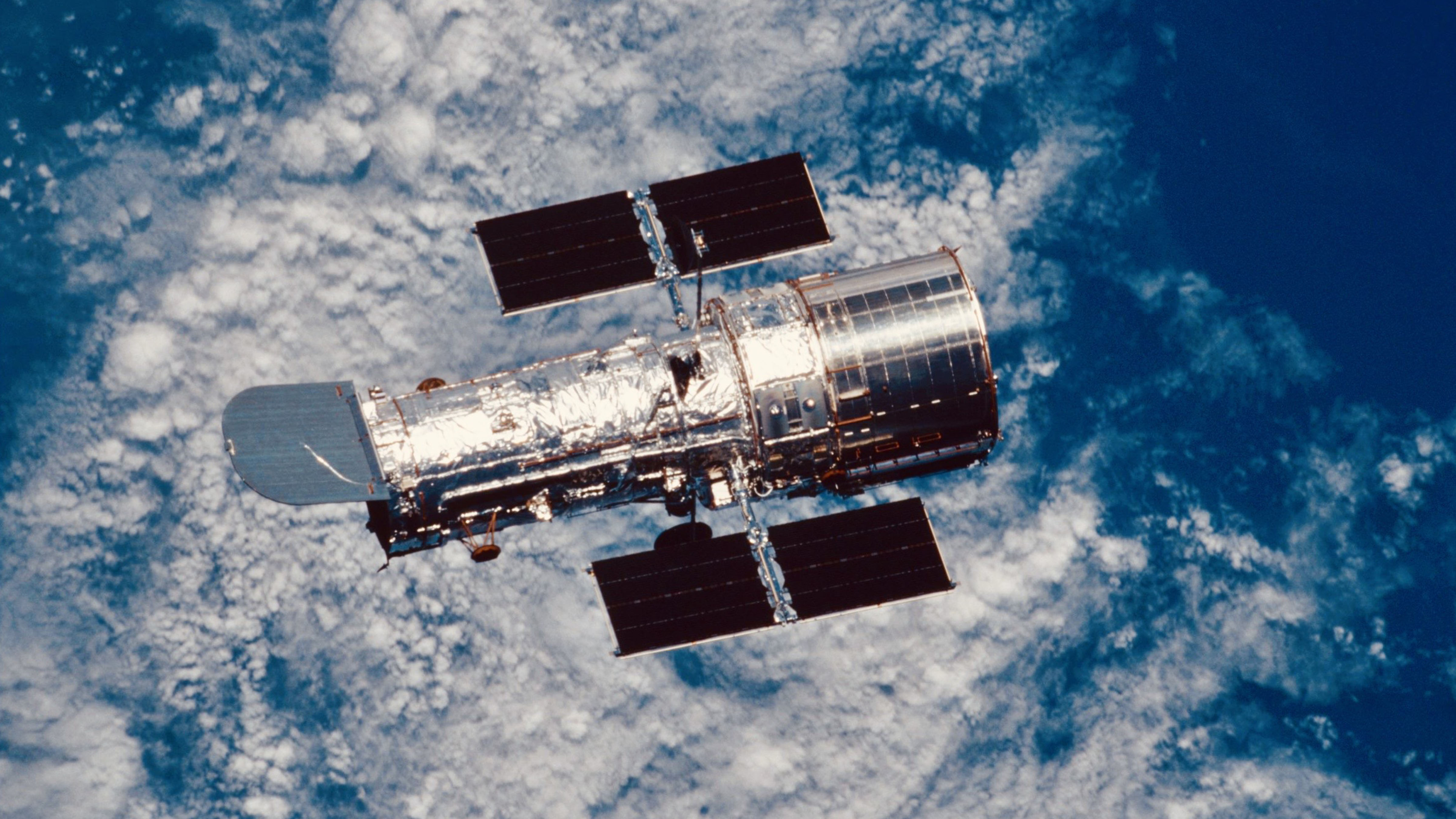 Hubble Space Telescope orbiting Earth with solar panels extended above clouds and atmosphere