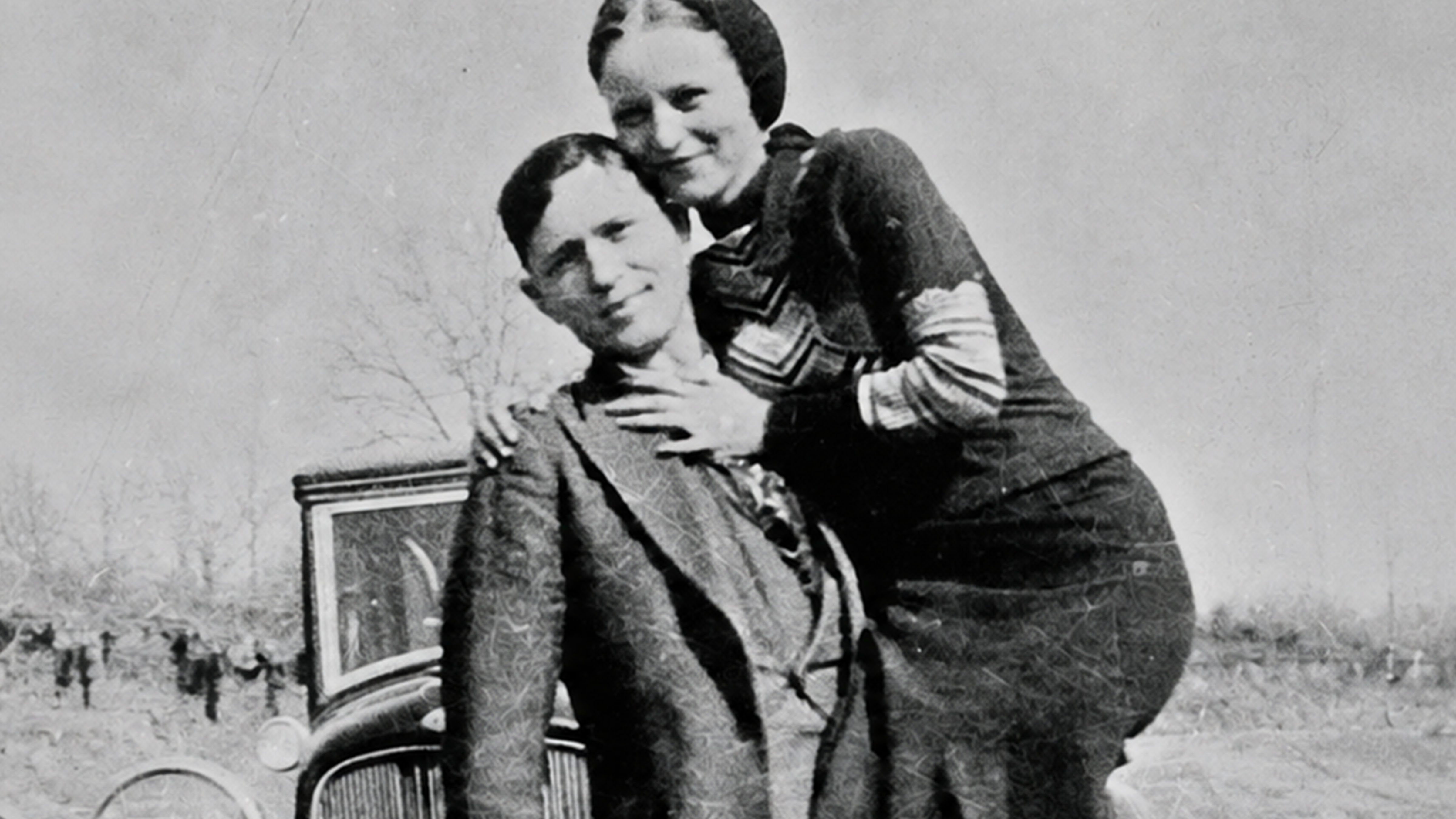 Historic black-and-white photograph of Bonnie Parker and Clyde Barrow standing together beside a car during the early 1930s.