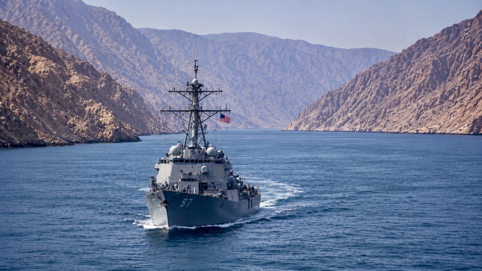 A U.S. Navy guided-missile destroyer sails through a narrow mountain-lined waterway, illustrating naval operations in the strategically important Strait of Hormuz.
