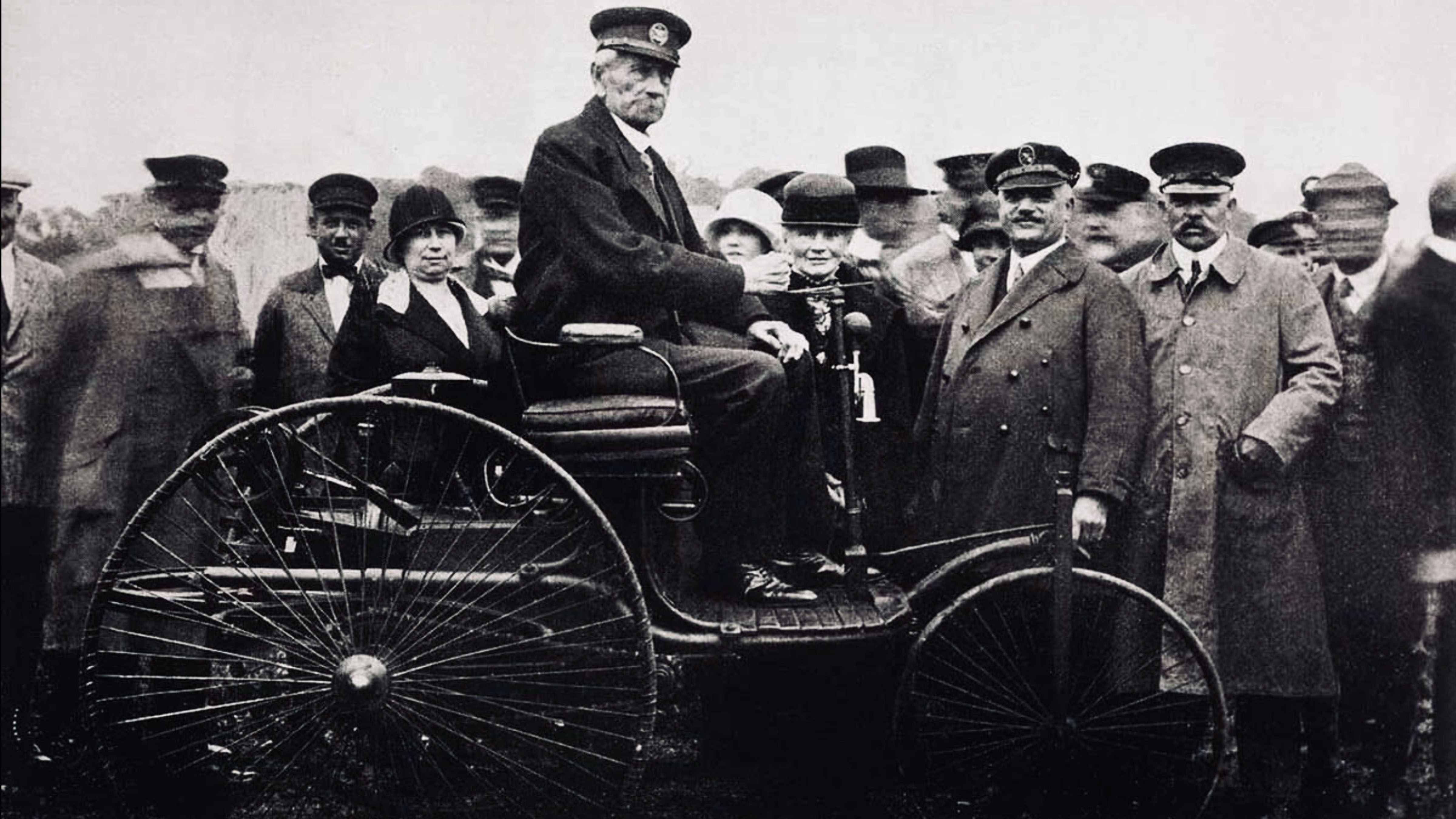 Karl Benz seated in one of his early automobiles as a crowd gathers to witness one of the pioneering demonstrations that helped launch the modern automobile era.
