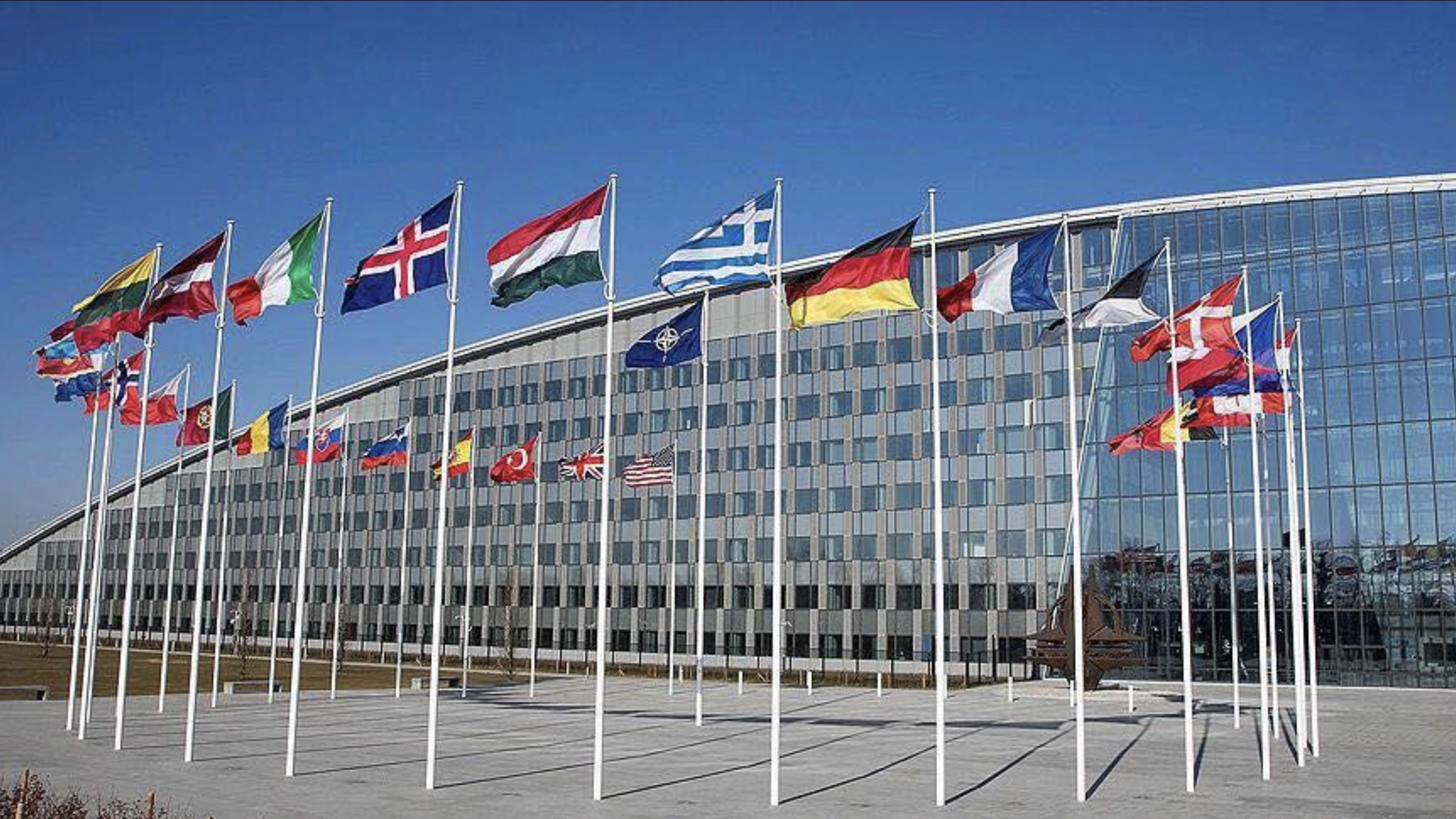 Flags of NATO member countries displayed outside NATO Headquarters in Brussels, Belgium, with the alliance’s modern headquarters building in the background.