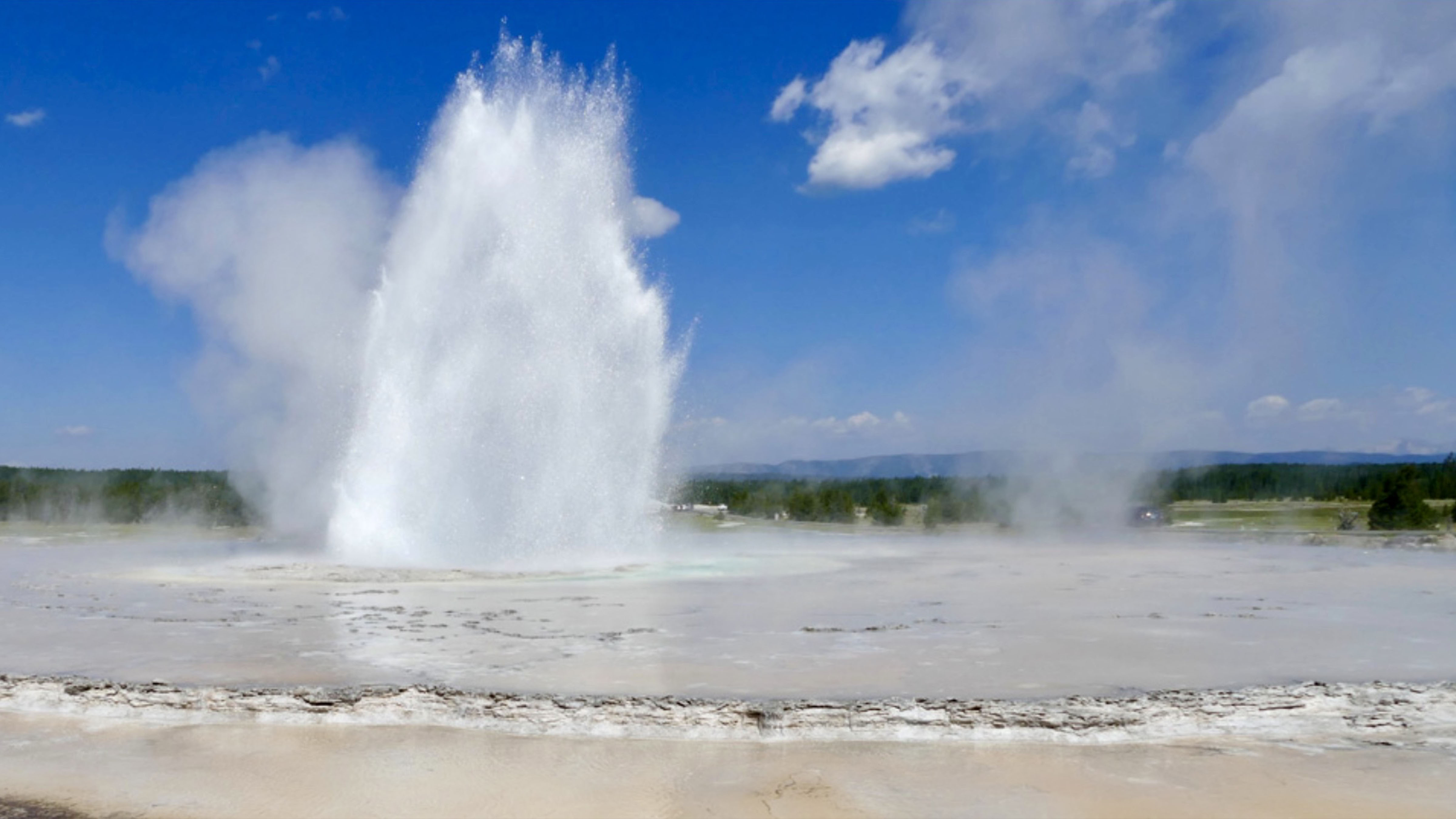 Old Faithful geyser erupts in Yellowstone National Park, sending a towering column of steam and hot water into the clear blue sky.